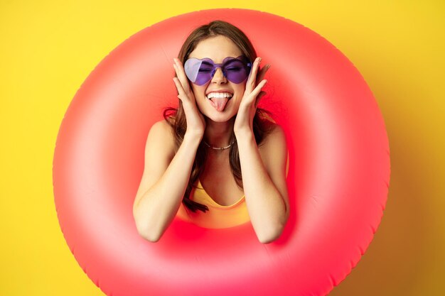 Feche o retrato de uma jovem entusiasmada dentro do anel de natação rosa, rindo e sorrindo, aproveitando as férias na praia, férias de verão, fundo amarelo
