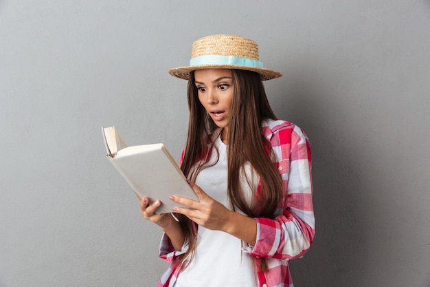 Feche o retrato de uma jovem espantada no chapéu de palha, lendo um livro