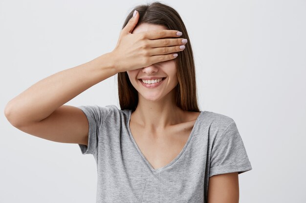 Feche o retrato de uma jovem mulher bonita e alegre alegre caucasiana com cabelos longos escuros na camiseta cinza, sorrindo com dentes, olhos de roupas com a mão, indo ver seu aniversário presente em alguns momentos.