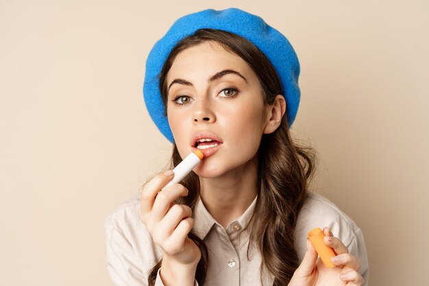 Feche o retrato de uma jovem mulher bonita olhando no espelho e consertando a maquiagem, coloque o batom, de pé sobre fundo bege. Conceito de beleza de cosméticos e mulheres