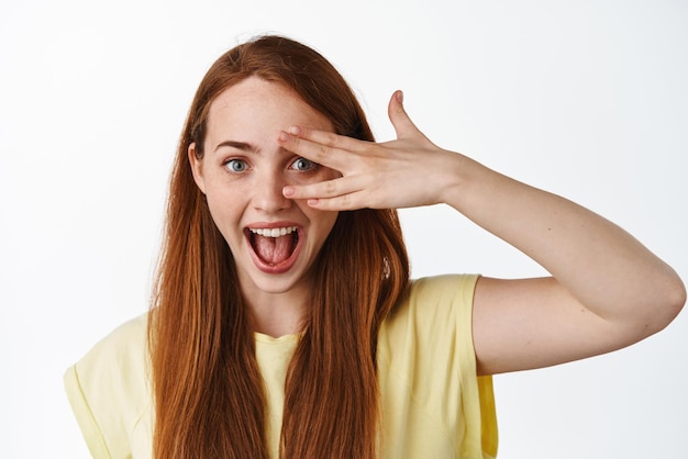 Feche o retrato de uma jovem ruiva atraente mostra uma emoção genuína e feliz entre os dedos e mostrando um fundo branco de pele facial limpa pura