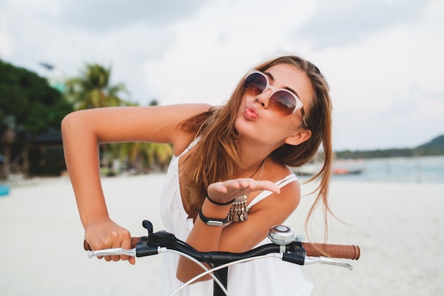 Feche o retrato de uma jovem sorridente em vestido branco andando em uma praia tropical com óculos de sol de bicicleta, viajando nas férias de verão na Tailândia