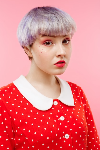 Feche o retrato de uma linda garota dollish com cabelo violeta leve curto, vestido vermelho sobre parede rosa