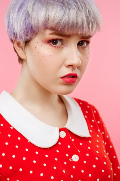 Feche o retrato de uma linda garota dollish com cabelo violeta leve curto, vestido vermelho sobre parede rosa