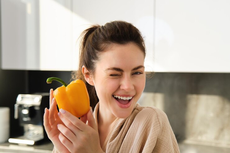 Foto grátis feche o retrato de uma linda garota piscando mostrando pimentão amarelo em pé na cozinha fazendo vegetariano