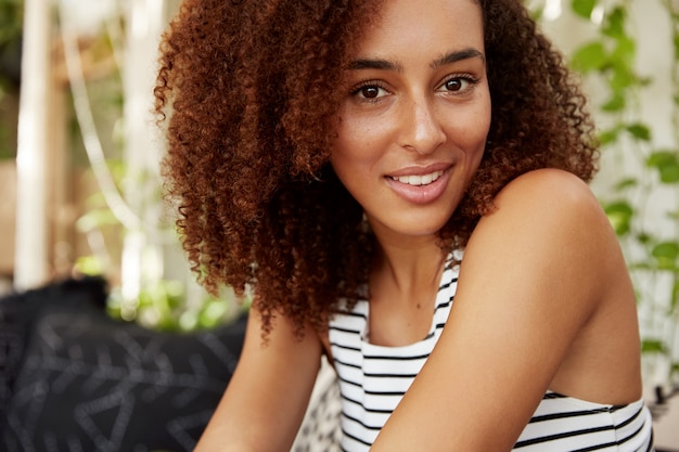 Feche o retrato de uma linda mulher de pele escura com penteado Afro tem aparência alegre e confiante, passa o tempo livre no refeitório. Jovem afro-americana bonita recriar depois do trabalho