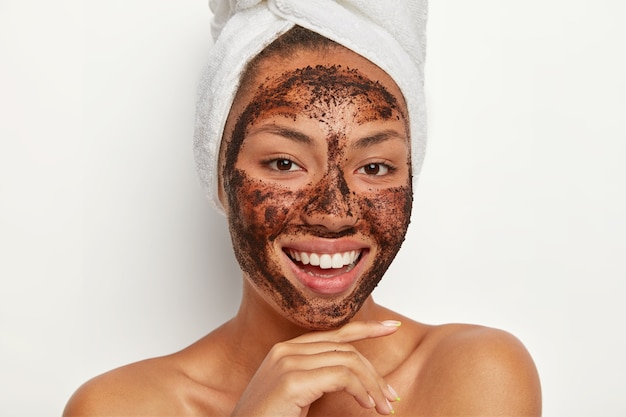 Foto grátis feche o retrato de uma mulher afro feliz que toca o queixo suavemente, sorri amplamente, mostra os dentes brancos, limpa o rosto, aplica uma máscara de café, usa uma toalha enrolada no cabelo molhado depois do banho. cuidados com a pele