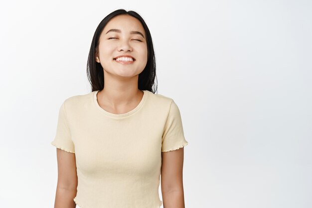 Feche o retrato de uma mulher asiática despreocupada sorrindo com os olhos fechados sonhando acordado imaginando algo de pé na camiseta amarela sobre fundo branco