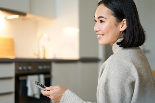 Feche o retrato de uma mulher asiática sorridente assistindo televisão sentada no sofá com controle remoto e switchi