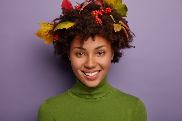 Feche o retrato de uma mulher atraente e cacheada se sentindo satisfeita, mostra dentes brancos perfeitos, sorri amplamente, tem um corte de cabelo estiloso com folhas outonais, expressa emoções positivas