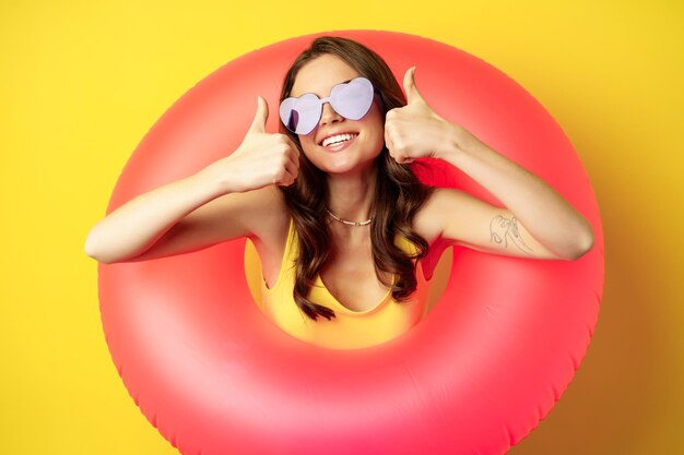 Feche o retrato de uma mulher atraente feliz em óculos de sol, usando anel de natação rosa, sorrindo e mostrando os polegares para cima, de pé sobre fundo amarelo