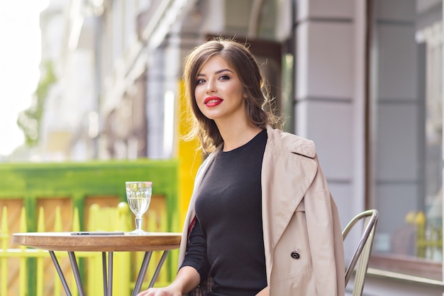 Feche o retrato de uma mulher bonita e charmosa com lábios vermelhos, usando um vestido preto e um casaco bege, descansando em um refeitório de verão com um copo d'água