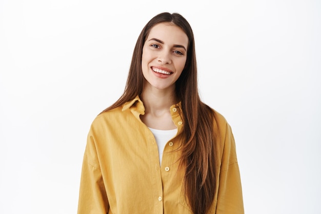 Feche o retrato de uma mulher caucasiana feliz com longos cabelos naturais nu maquiagem sorrindo e inclinando a cabeça parecendo amigável e positivo em pé sobre fundo branco