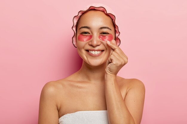 Feche o retrato de uma mulher coreana feliz fazendo um sinal, cruza o polegar e o indicador, manda amor, usa touca de banho rosa, faz tratamentos de cosmetologia sob os olhos, aplica adesivos de colágeno para hidratar