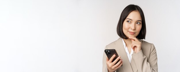 Feche o retrato de uma mulher corporativa coreana de terno usando telefone celular e sorrindo segurando smartphone em pé sobre fundo branco