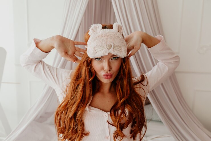 Foto grátis feche o retrato de uma mulher engraçada com cabelo ruivo comprido e ondulado e máscara de dormir fazendo caretas na cama