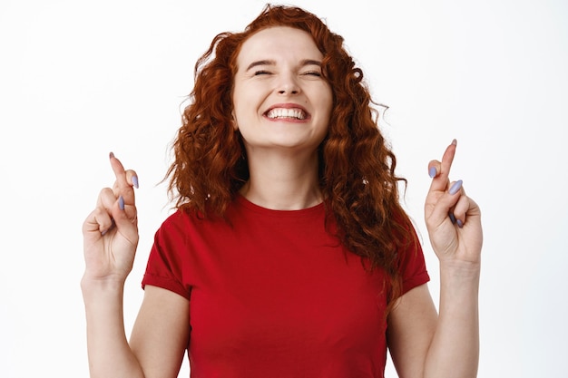 Foto grátis feche o retrato de uma mulher esperançosa e animada, sorrindo, em pé com os dedos cruzados e fazendo pedidos, rezando para que o sonho se tornasse realidade, implorando, em pé contra uma parede branca