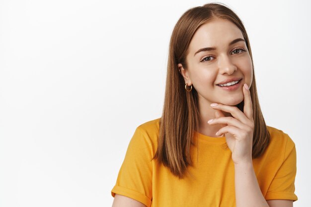 Feche o retrato de uma mulher loira sorridente, parecendo intrigado, sorrindo, pensativo, sugestão interessante, parado satisfeito em uma camiseta amarela contra uma parede branca