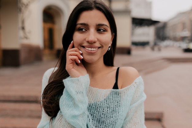 Feche o retrato de uma mulher morena tocando sua bochecha e sorrindo, vestindo uma camisa azul