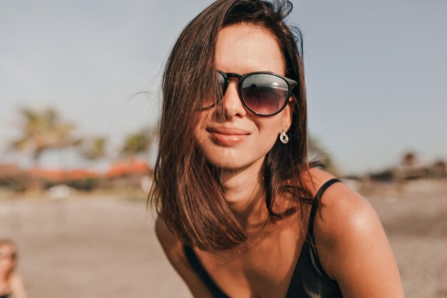 Feche o retrato de uma mulher muito feliz com cabelo escuro, usando óculos escuros pretos, posando durante a sessão de fotos na praia perto do oceano