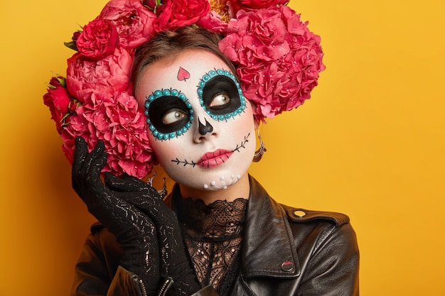 Feche o retrato de uma mulher pensativa, usa maquiagem criativa, pintou o sorriso, coroa de flores ao redor da cabeça, parece pensativamente longe.