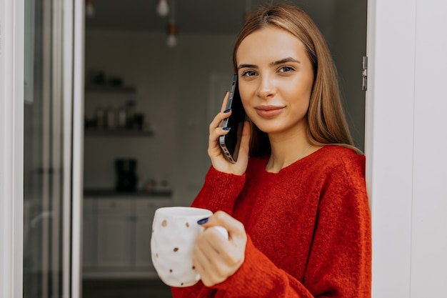 Feche o retrato de uma senhora alegre de suéter vermelho segurando uma xícara e falando ao telefone na cozinha