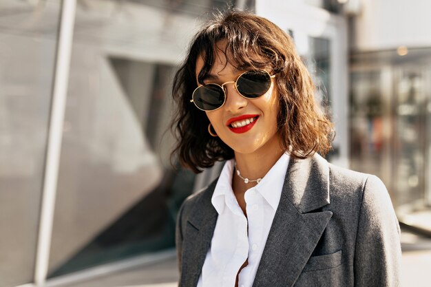 Feche o retrato de uma senhora bonita sorridente com penteado curto e batom vermelho, vestindo roupas casuais, posando para a câmera no fundo da cidade