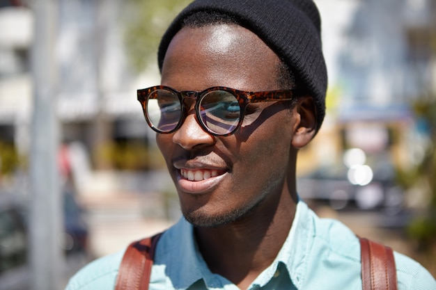 Feche o retrato de verão de bonito alegre jovem estudante europeu preto em óculos da moda e chapéu, sorrindo alegremente, aproveitando a manhã ensolarada