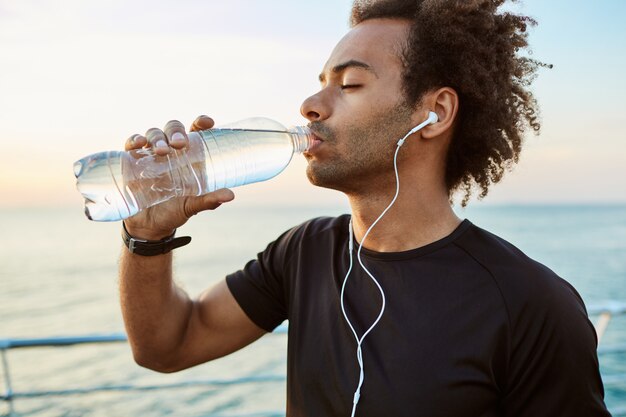 Feche o retrato do atleta afro-americano apto a beber água de uma garrafa de plástico com fones de ouvido. Refrescando-se com água e vestindo camiseta preta