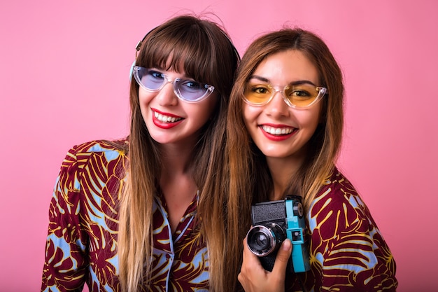 Feche o retrato do estúdio de estilo de vida de duas lindas amigas usando roupas e óculos escuros estampados, segurando uma câmera vintage