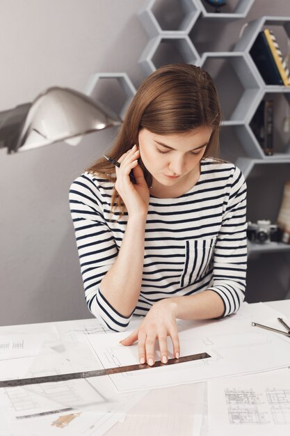 Feche o retrato do freelancer bonito jovem arquiteto feminino com cabelos longos escuros na camisa listrada, sentado à mesa branca no espaço de coworking, olhando através de plantas que ela já fez, pensando