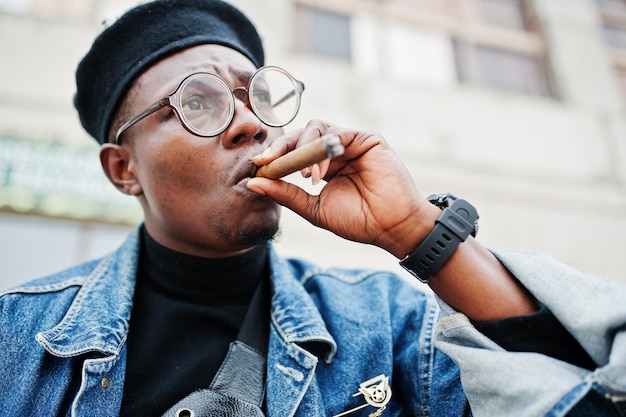 Feche o retrato do homem afro-americano na boina de jaqueta jeans e óculos fumando charuto