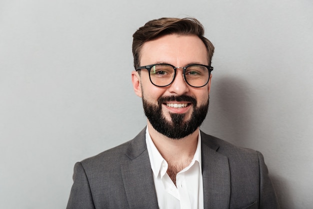 Feche o retrato do homem com barba caucasiano de óculos, olhando para a câmera com um sorriso sincero, isolado sobre cinza