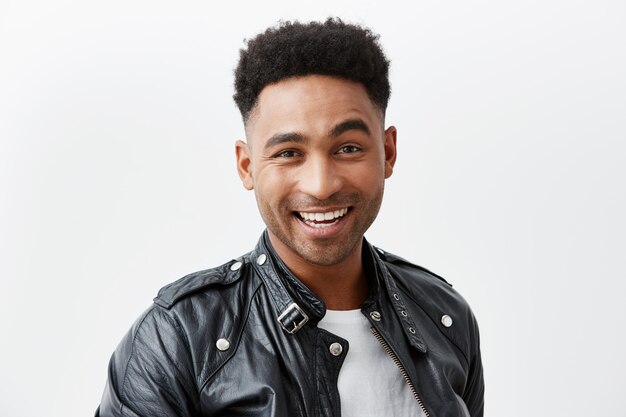 Feche o retrato do jovem americano alegre de pele escura com cabelos cacheados em camiseta branca e jaqueta de couro, sorrindo brilhantemente, olhando na câmera com a expressão do rosto feliz e animado.