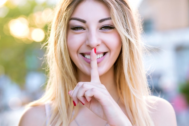 Feche o retrato ensolarado positivo de mulher jovem bonita loira feliz colocar o dedo na cara e dizer shhh, conceito secreto, maquiagem fresca natural.