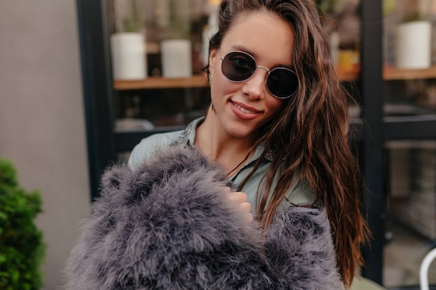 Feche o retrato externo de uma jovem encantadora usando pele e óculos elegantes, posando para a câmera