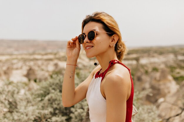 Foto grátis feche o retrato externo de uma mulher espetacular tocando os óculos e olhando para o lado com um sorriso na paisagem montanhosa