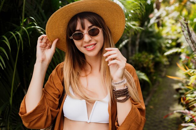 Feche o retrato na moda do verão de uma mulher morena com chapéu de palha, posando em folhas de palmeira tropical em Bali.