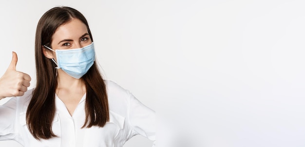 Foto grátis feche o rosto do retrato da mulher com máscara médica sorrindo mostrando os polegares para cima em pé sobre fundo branco