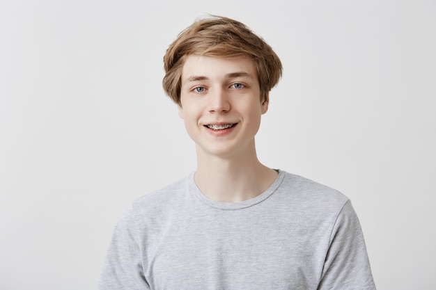 Felicidade e expressões humanas positivas. Foto de estúdio de amigável jovem estudante do sexo masculino com cabelo loiro e olhos azuis, rindo de boa piada, sorrindo com aparelho, olhando com sorriso despreocupado