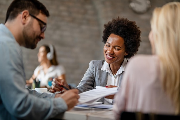 Feliz agente de seguros afro-americano analisando os termos do contrato com um casal e apontando para o local que eles precisam assinar no escritório