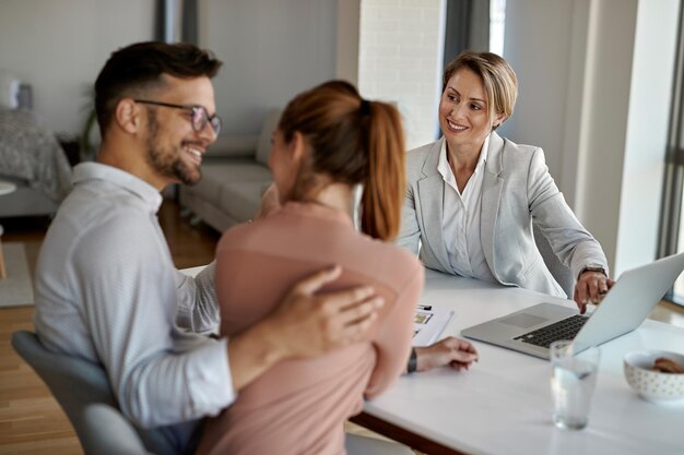 Feliz agente imobiliário usando laptop enquanto conversa com um casal durante a reunião no escritório