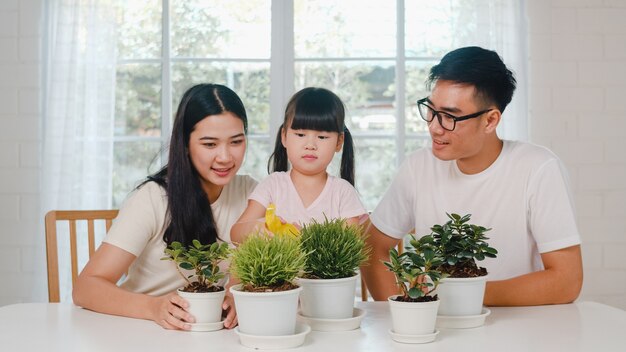 Feliz alegre família asiática pai, mãe e filha regando plantas em jardinagem perto de janela em casa