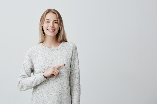 Feliz alegre jovem loira com cabelos tingidos, sorrindo amplamente apontando com o dedo indicador na parede com espaço de cópia