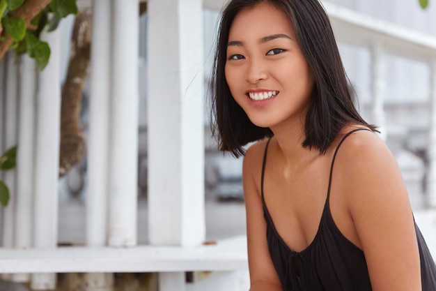 Foto grátis feliz, alegre, linda mulher asiática com largo sorriso satisfeito, vestida casualmente, tem bom descanso em um lugar calmo, expressa emoções positivas