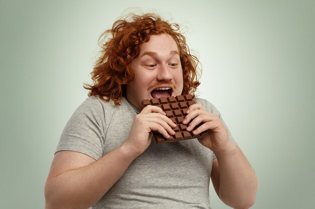 Feliz animado jovem ruiva gordinha masculina abertura boca amplamente enquanto morde a barra de chocolate, sentindo-se impaciente. Engraçado homem caucasiano de camiseta cinza, consumindo alimentos pouco saudáveis, mas deliciosos