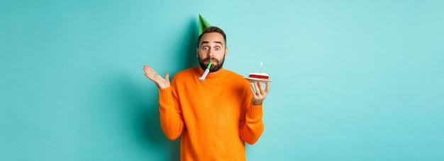 Foto grátis feliz aniversariante comemorando usando chapéu de festa soprando apito e segurando bolo de aniversário em pé novamente