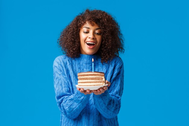 Feliz aniversariante sonhadora e esperançosa fazendo desejo. Mulher afro-americana atraente com corte de cabelo encaracolado, inale o ar para apagar a vela acesa no saboroso bolo de aniversário, parede azul de pé.