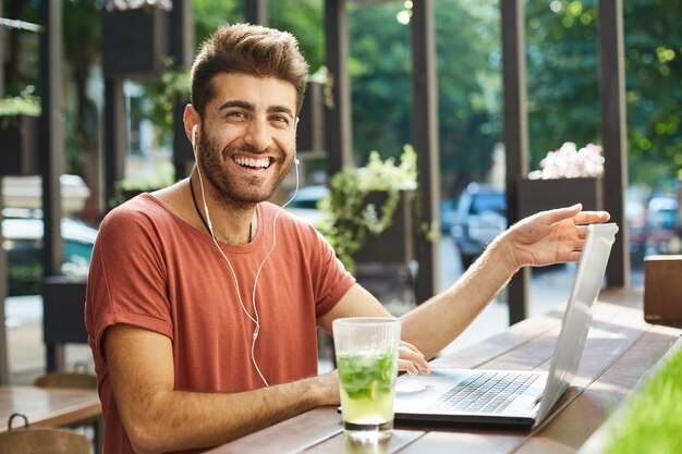 Feliz barbudo bonito usando fones de ouvido usando laptop em um café ao ar livre, sorrindo alegremente