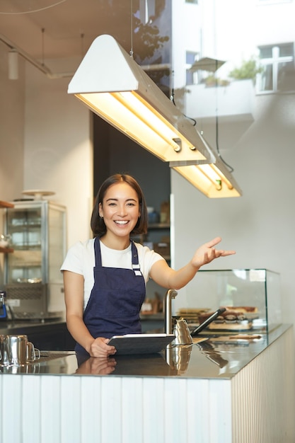 Foto grátis feliz barista asiática trabalha em ondas de cafeteria em pedidos de processamento de clientes atrás do balcão em